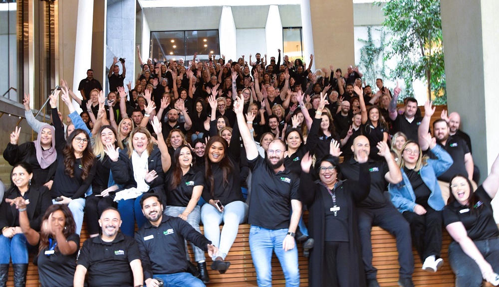 A large group of people in black polo shirts with Jobfind logo wave and smile at the camera.