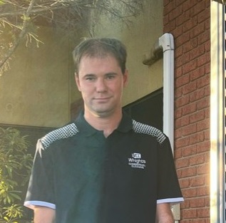 Young man with dark hair, dressed in a dark shirt and jeans, stands in front of a brick building.