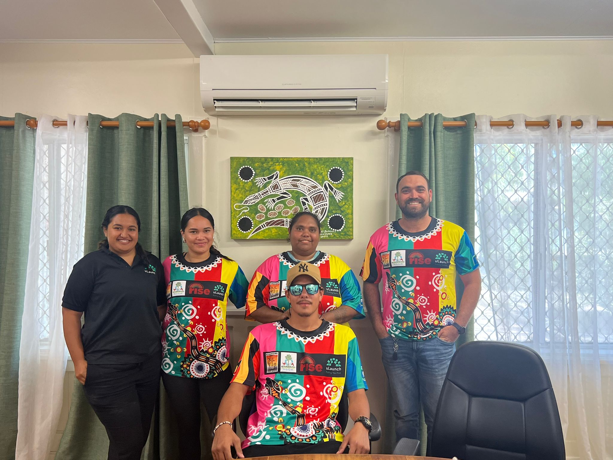 Five people in an office environment wear a brightly-coloured indigenous t-shirt that won the competition.