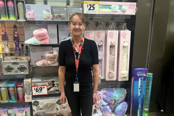 Kristie, dressed in black top and trousers, with lanyard and her ID, stands in front of shop shelves at work.